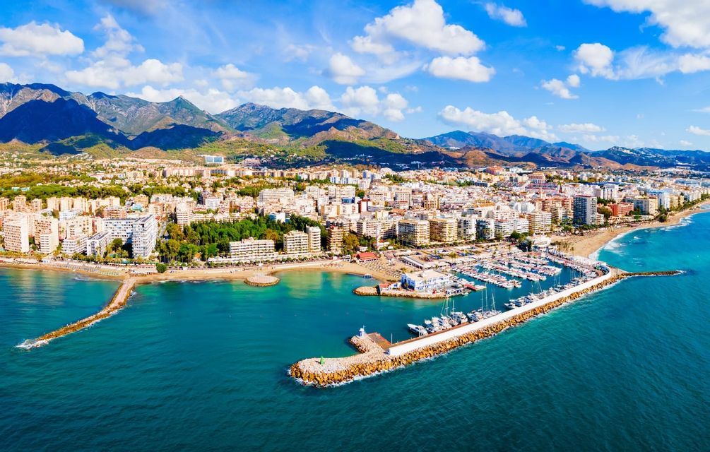 Vista aerea di una città costiera con un grande porto turistico, spiaggia sabbiosa e montagne sullo sfondo sotto un cielo blu e nuvoloso.
