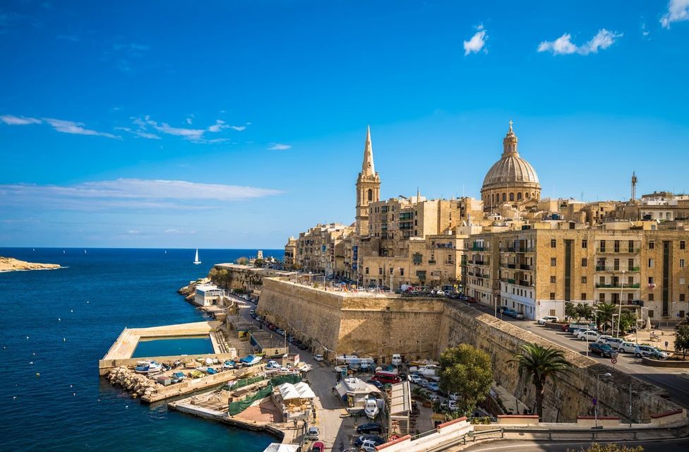 Una città costiera fortificata, con una cupola e un campanile ben visibili, si affaccia su un porto e sul mare blu profondo in una giornata di sole.