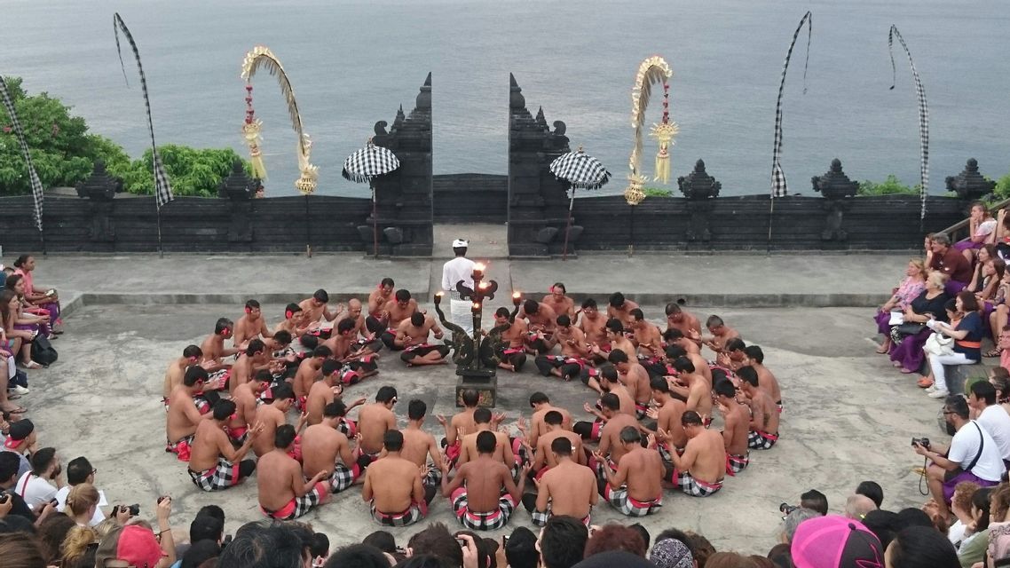 Il pubblico osserva uomini in pareo seduti in cerchio per una tradizionale danza del fuoco, in un luogo all'aperto in riva al mare.