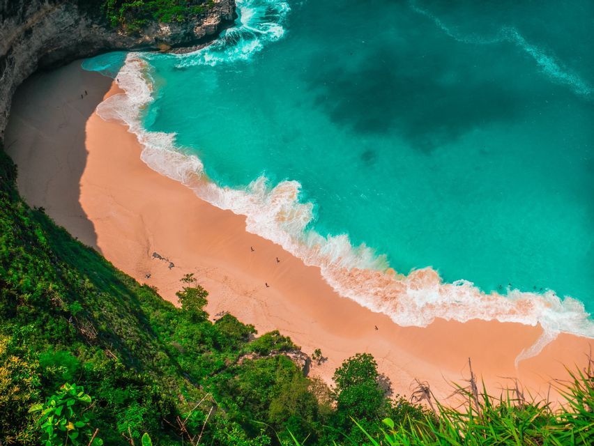 Una vista aerea di una spiaggia sabbiosa isolata incastonata contro una scogliera, con onde turchesi che si infrangono sulla riva e alcune persone visibili.