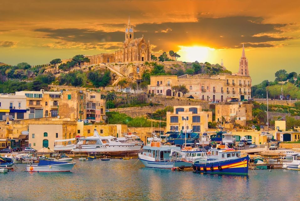 Eine Küstenstadt mit einer Kirche auf einem Hügel überblickt bei Sonnenuntergang einen Hafen voller Boote und Yachten.