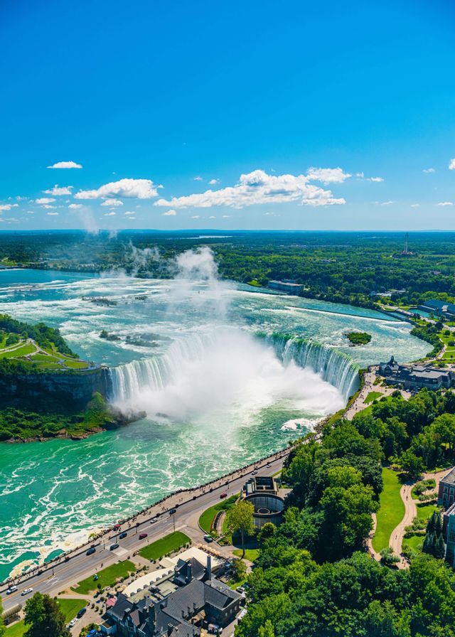 Una veduta aerea di una vasta cascata a ferro di cavallo che si riversa in un fiume turchese, con una strada e una città immerse nel lussureggiante paesaggio verde.