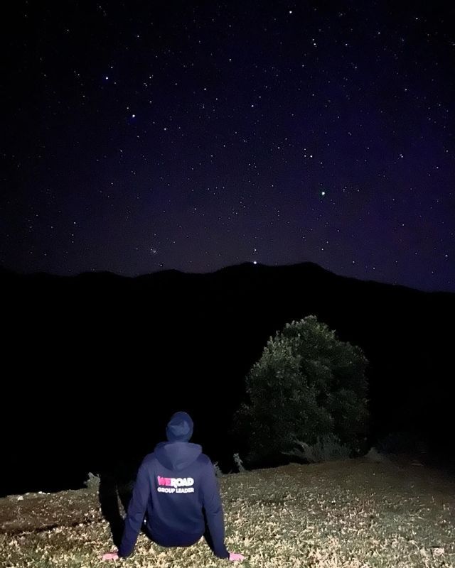 Un capogruppo WeRoad siede su una collina di notte, guardando un cielo stellato sopra la silhouette di una montagna.