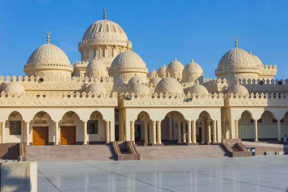 Una moschea color crema riccamente decorata, con cupole e archi multipli, si erge sotto un cielo azzurro limpido, preceduta da un vasto cortile.