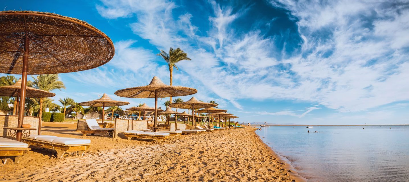 Ombrelloni di paglia e sedie a sdraio fiancheggiano una spiaggia sabbiosa accanto al mare calmo sotto un cielo azzurro brillante, parzialmente nuvoloso.