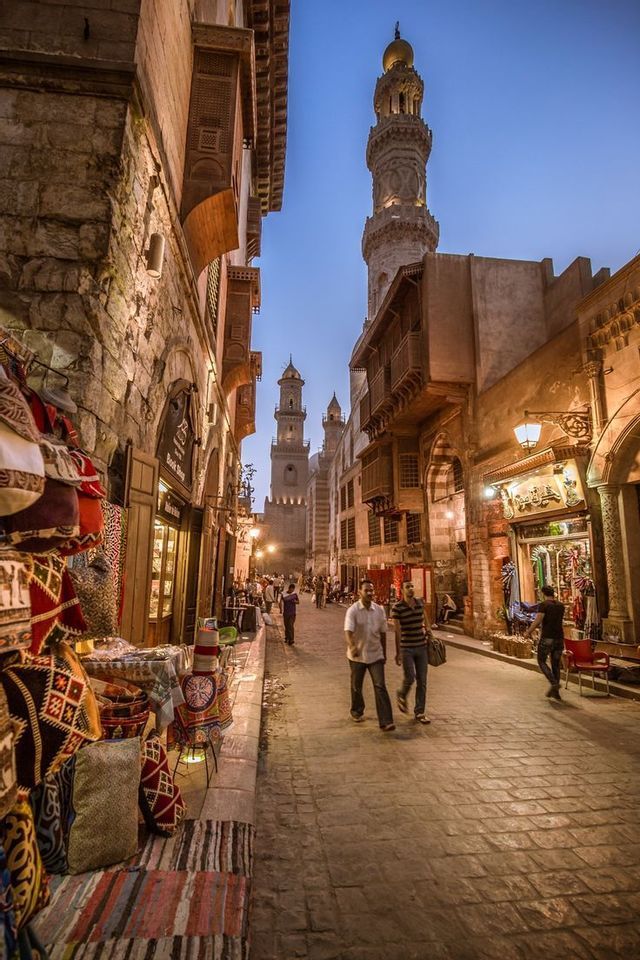 La gente passeggia lungo una stretta via di mercato acciottolata al tramonto, con negozi illuminati e minareti storici sullo sfondo.