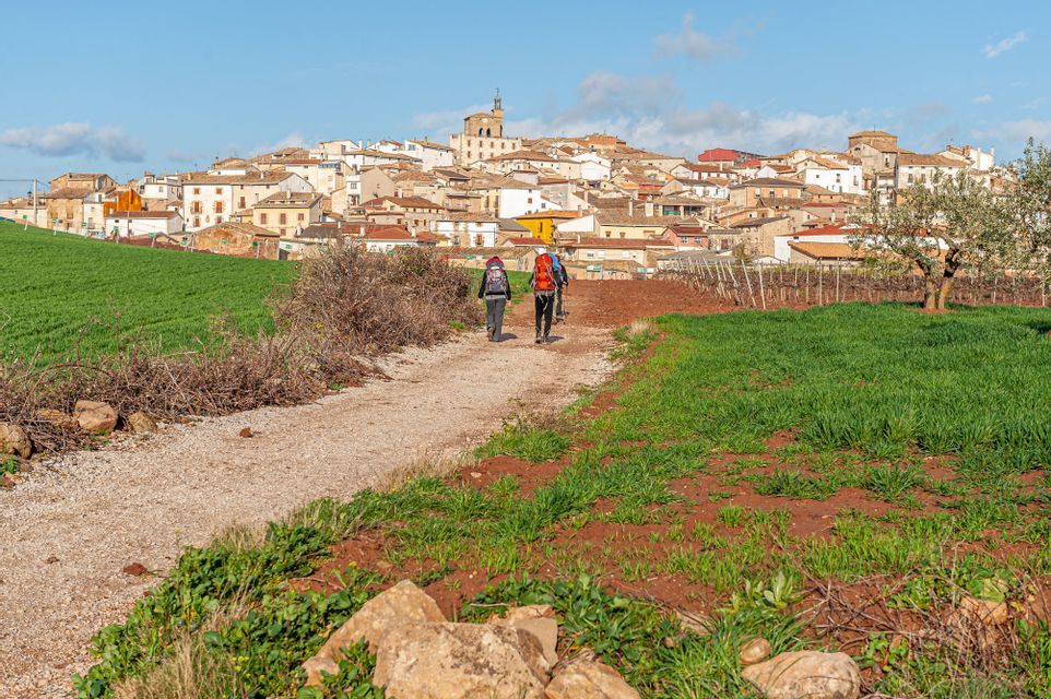 Due persone con zaini da trekking percorrono un sentiero di ghiaia attraverso campi verdi verso un borgo in collina.