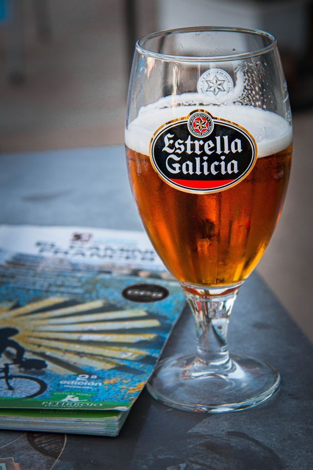 Nahaufnahme eines Glases Estrella Galicia Bier mit Schaumkrone, auf einem dunklen Tisch neben einer Zeitschrift stehend.