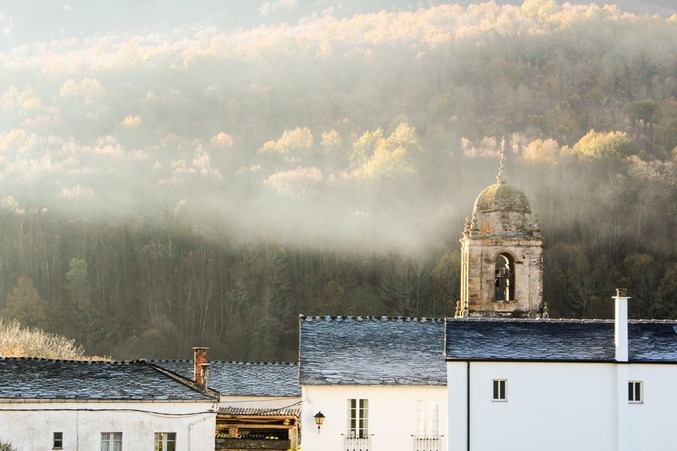 Un campanile in pietra si innalza sopra le case bianche del villaggio, con tetti in ardesia, su un fianco di collina nebbioso e boscoso.