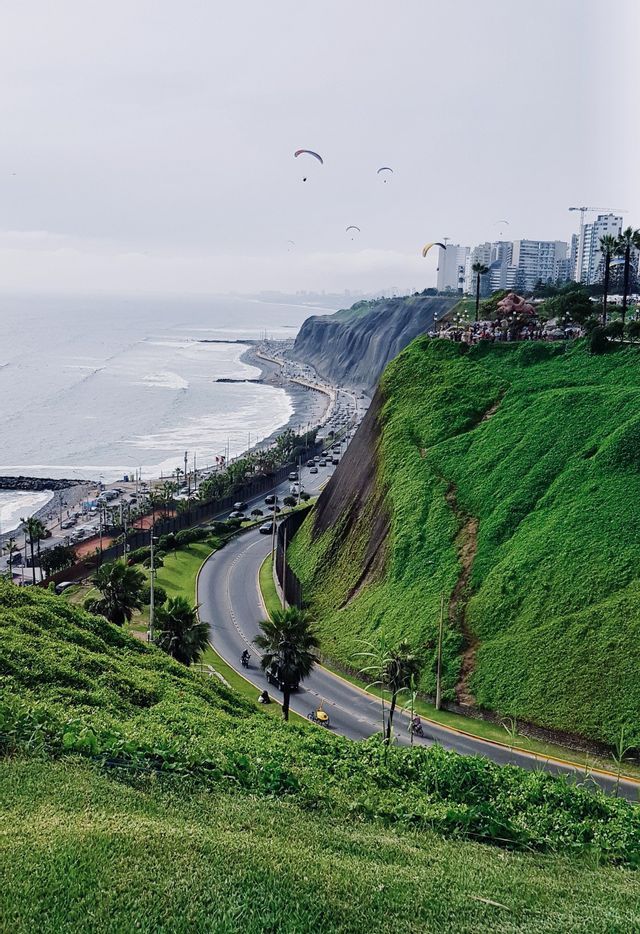 Paraglider gleiten über eine kurvige Küstenstraße und grüne Klippen am Meer, mit Stadtgebäuden im Hintergrund.
