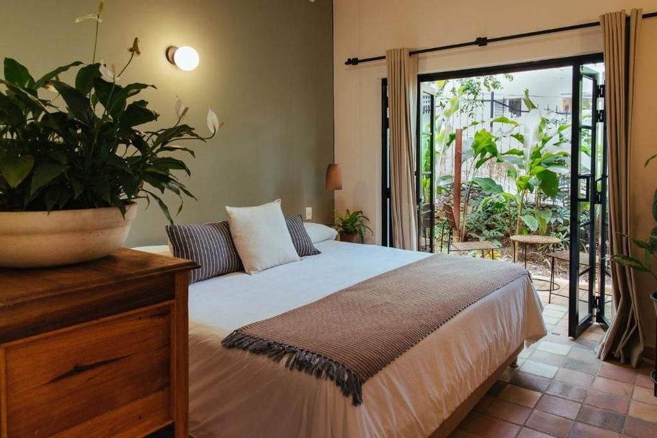Interior of a bedroom with a double bed, a large potted plant, and open doors leading to a green patio.