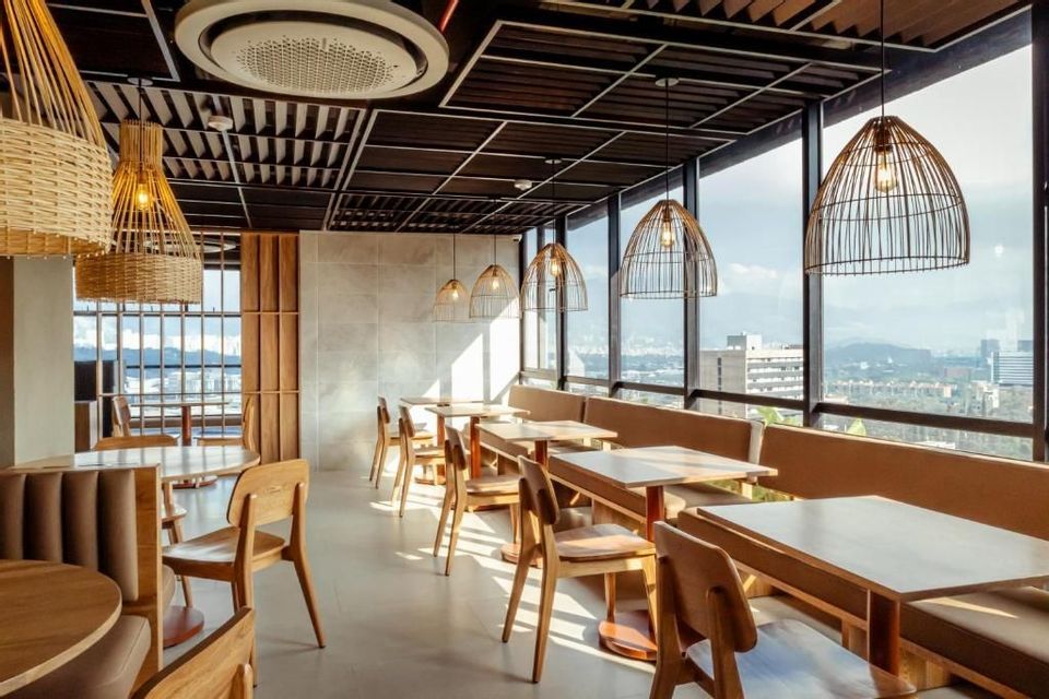 The sunlit interior of a modern restaurant with wooden furniture, wicker pendant lights, and large windows overlooking a cityscape.