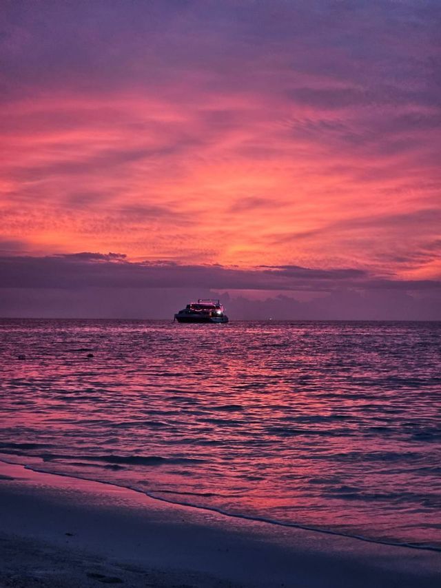 Una barca illuminata galleggia sul mare sotto un cielo al tramonto rosa e viola, con una costa in primo piano.