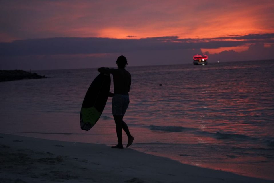 Una persona in silhouette con una tavola da surf cammina lungo la riva al tramonto, con una barca sull'acqua in lontananza.
