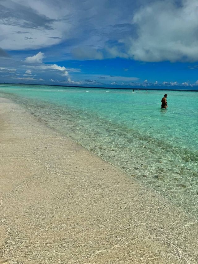 Un uomo in piedi nell'acqua bassa e trasparente color turchese, con una spiaggia sabbiosa in primo piano sotto un cielo nuvoloso.