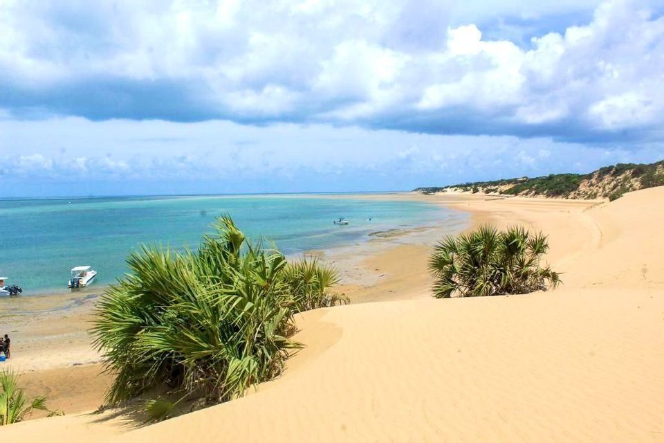 Blick von einer Sanddüne mit grünen Büschen auf eine türkisfarbene Bucht mit am Strand ankernden Booten unter bewölktem Himmel.