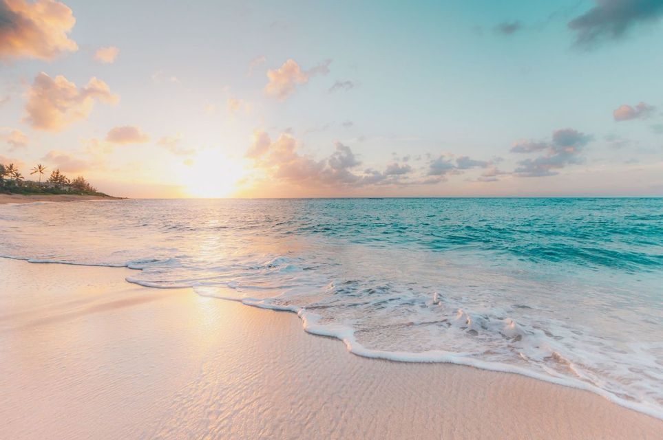 Il sole tramonta su un oceano calmo e turchese, con onde delicate che si infrangono su una spiaggia sabbiosa sotto un cielo pastello.