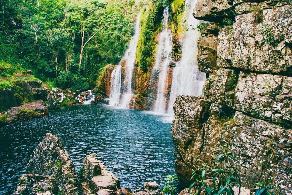 Ein Wasserfall stürzt eine moosbewachsene, terrassenförmige Felsklippe hinab in einen tiefblauen Pool, umgeben von einem üppig grünen Wald.