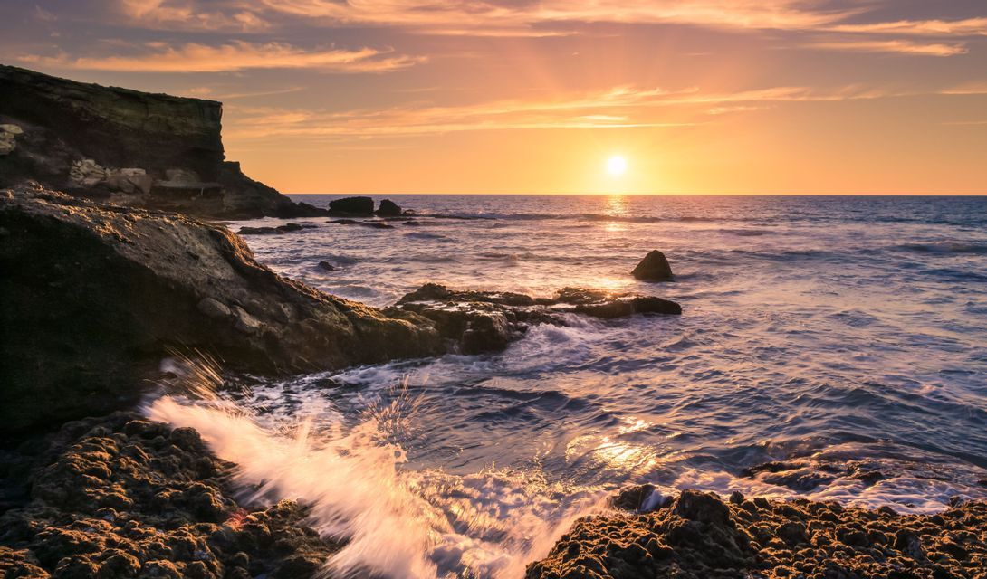 Meereswellen brechen sich an einer felsigen Küste unter einem goldenen Sonnenuntergang, während die Sonne tief am Horizont steht.
