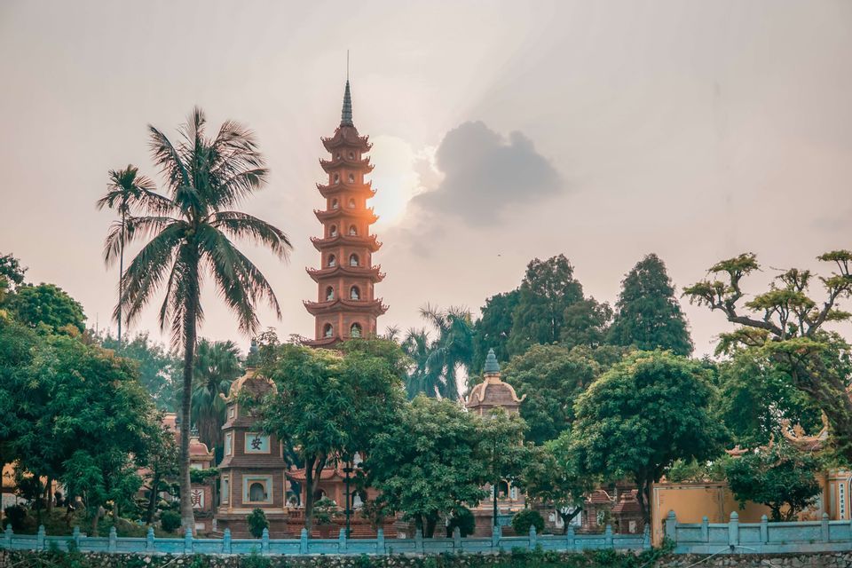 Una pagoda a più livelli si erge tra alberi verdi lussureggianti e una palma mentre il sole tramonta dietro di essa.