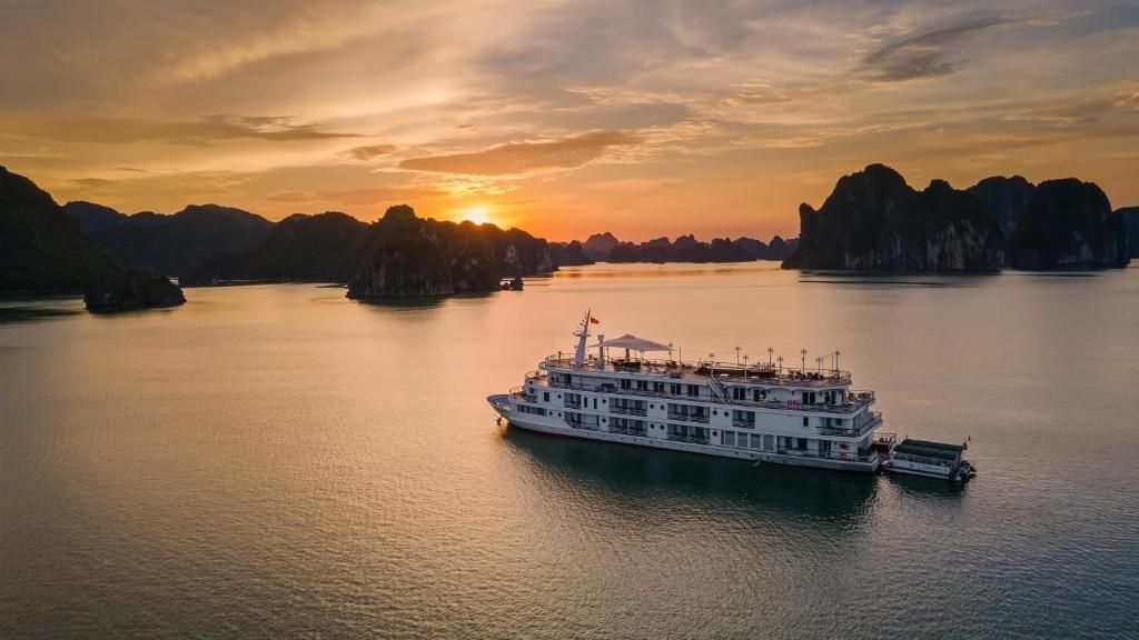 Una nave da crociera bianca galleggia su acque calme, circondata da isole rocciose mentre il sole tramonta all'orizzonte.