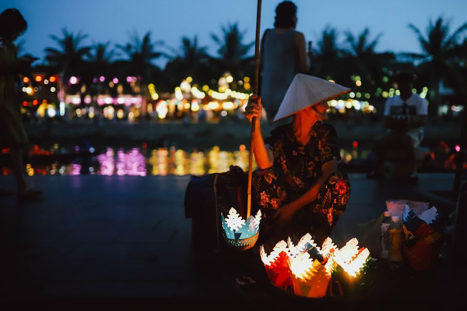 Un venditore con un cappello conico vende lanterne di carta colorate e illuminate sulla riva di un fiume al tramonto, con le luci della città sullo sfondo.