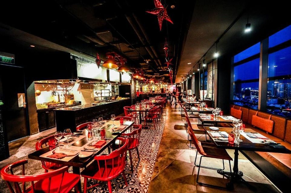 Interior de restaurante cálidamente iluminado con sillas rojas y mesas puestas junto a grandes ventanales con vistas al horizonte de una ciudad por la noche.