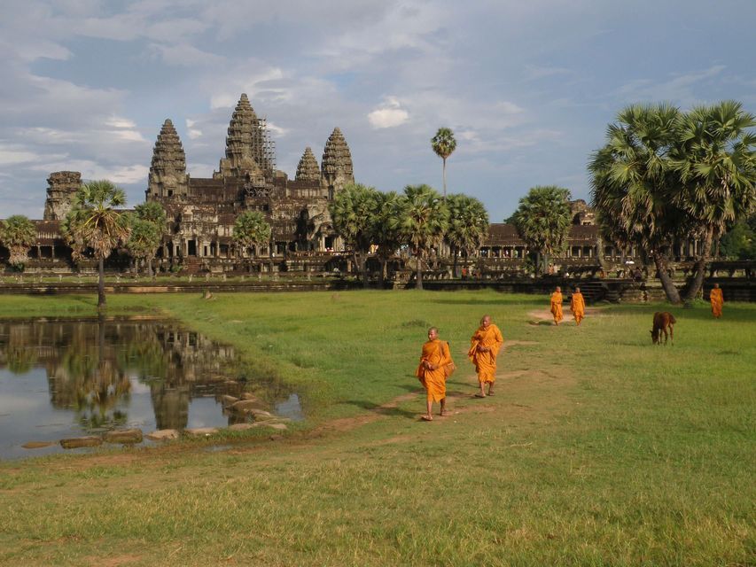 Des moines aux robes orange marchent sur un chemin devant un ancien temple de pierre, qui se reflète dans un étang proche.