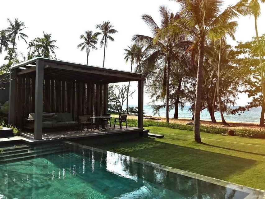 Una piscina e una cabana in legno su un prato erboso, accanto a una spiaggia sabbiosa con palme e l'oceano.