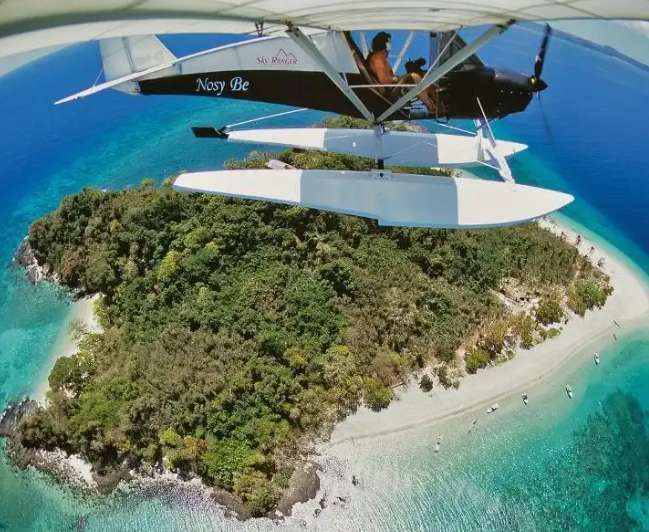 Una vista fisheye da un idrovolante ultraleggero in volo sopra un'isola tropicale lussureggiante con una spiaggia di sabbia bianca e acqua turchese.
