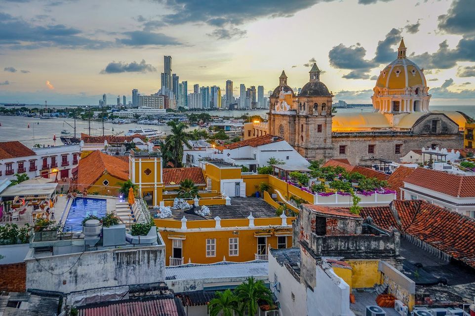 Veduta urbana di edifici storici con tetti in terracotta e una cattedrale, con uno skyline moderno sull'acqua al tramonto.