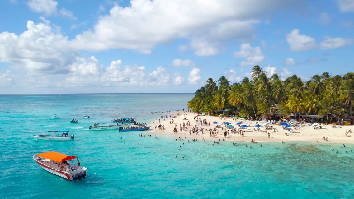 Una vista aerea di una spiaggia tropicale affollata con acqua turchese, numerose imbarcazioni e persone che nuotano e prendono il sole sulla sabbia.