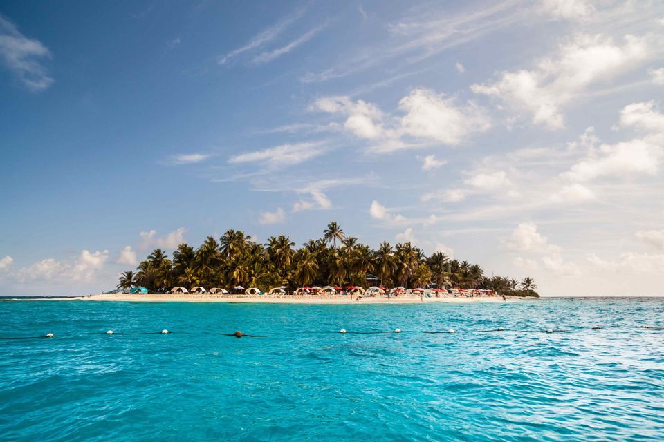 Un'isola tropicale con palme e tende colorate su una spiaggia sabbiosa, vista dal mare turchese sotto un cielo parzialmente nuvoloso.