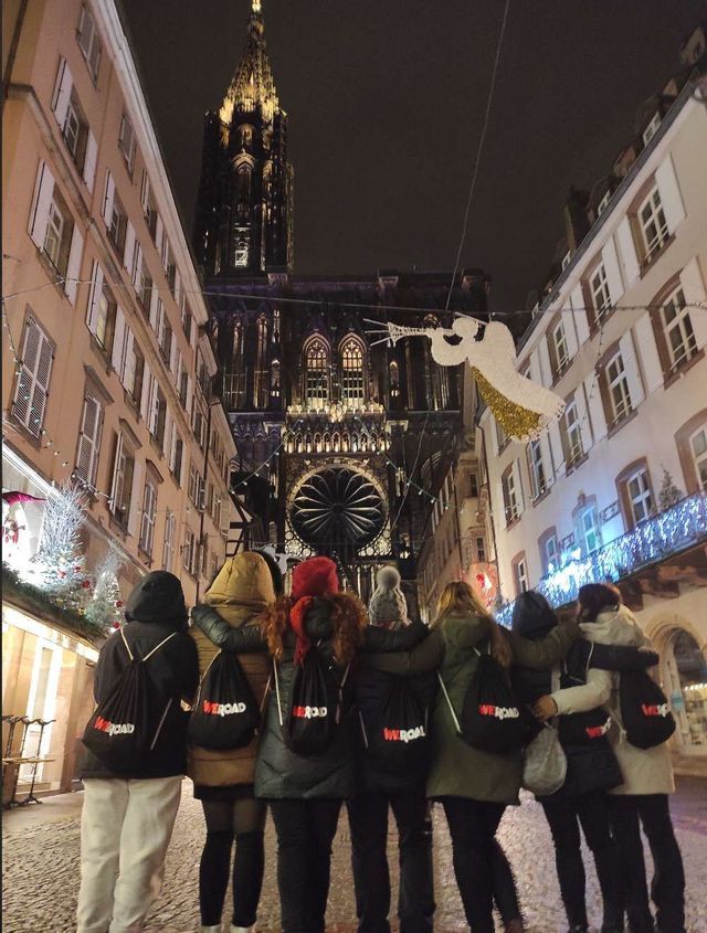 A WeRoad group trip seen from behind, standing arm-in-arm on a decorated cobblestone street at night, looking at a large illuminated cathedral.