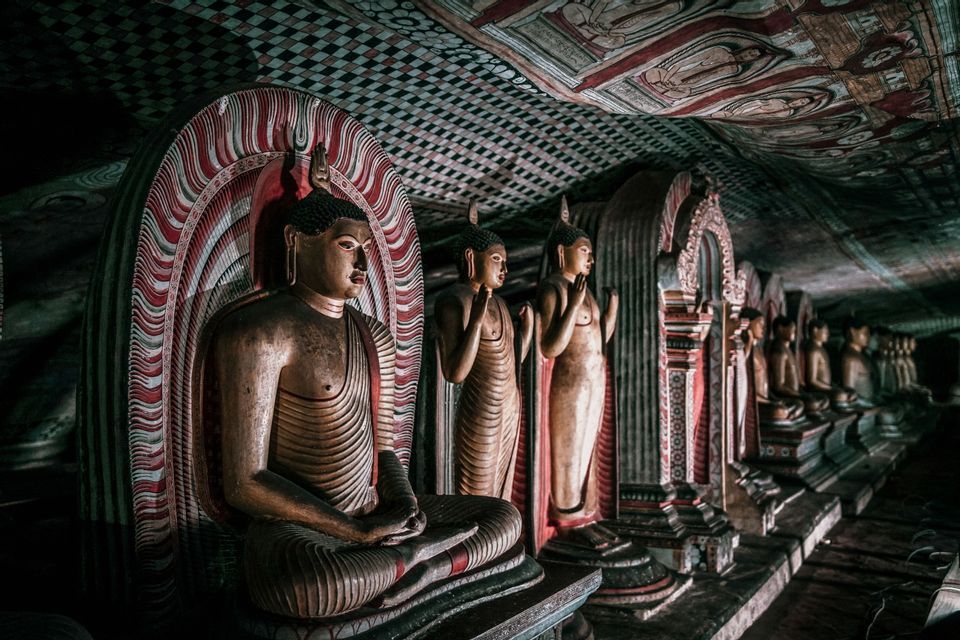 Une longue rangée de statues de Bouddha sculptées à l'intérieur d'un temple rupestre faiblement éclairé avec un plafond richement peint.
