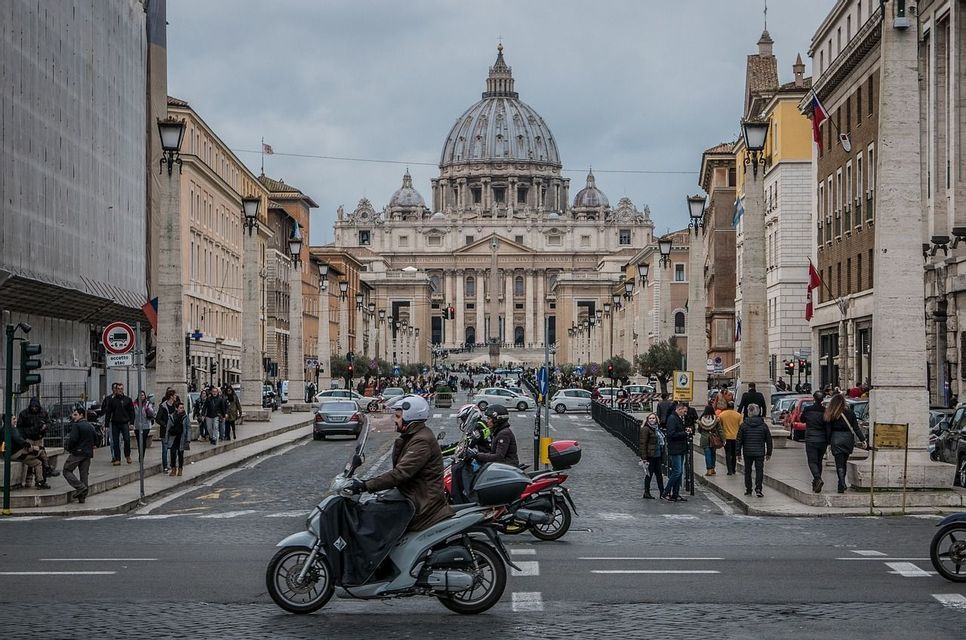 Una amplia calle adoquinada con tráfico, motos y peatones, que conduce a una gran basílica con cúpula bajo un cielo cubierto.