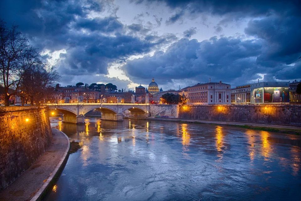 Vista de un paisaje urbano al anochecer mostrando un puente arqueado iluminado sobre un río, con una gran cúpula al fondo bajo un cielo nublado.