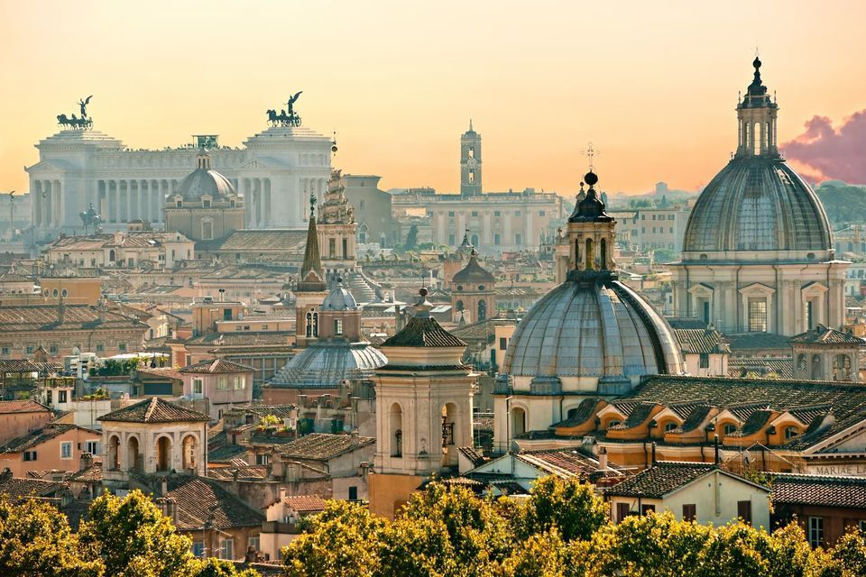 A cityscape of historic domes, monuments, and buildings with terracotta roofs seen from above under a hazy golden sky.
