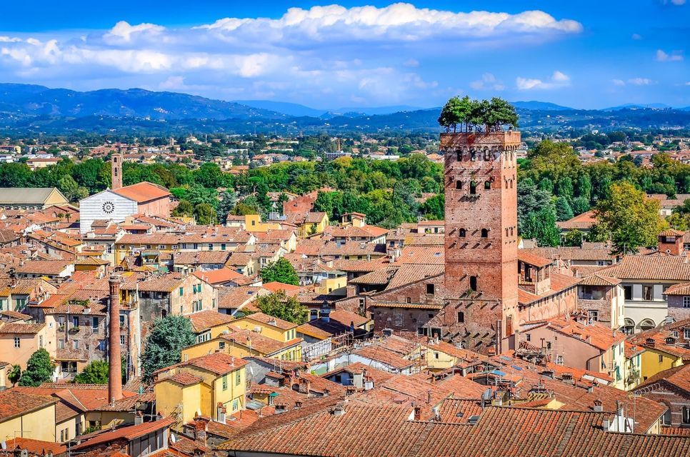 An aerial view of a city with terracotta-tiled roofs, featuring a prominent brick tower with trees growing on its summit.