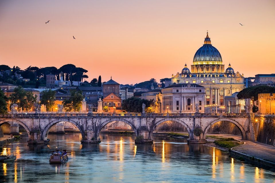A grand domed basilica and an arched stone bridge are illuminated over a river at sunset, with a tour boat in the water.