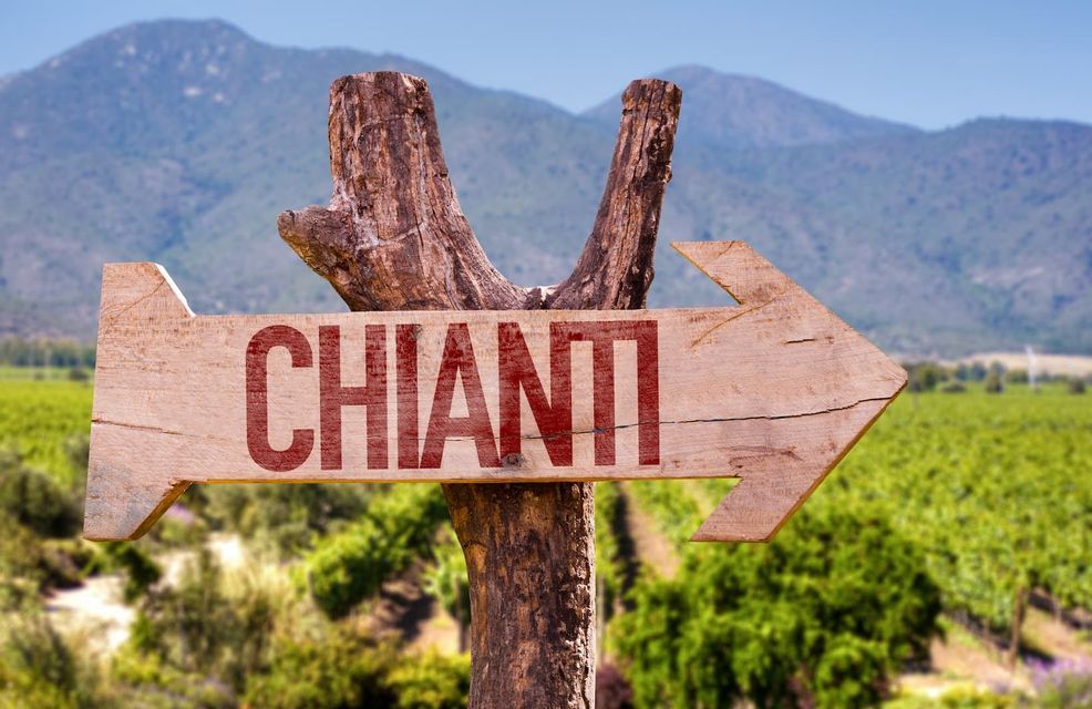 A wooden signpost shaped like an arrow points right with the word 'CHIANTI' in red, set against a view of vineyards and mountains.