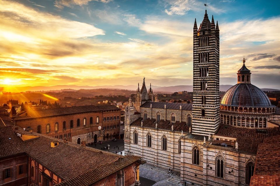 An aerial view of a cathedral with a striped bell tower and dome overlooking a historic city at sunset.