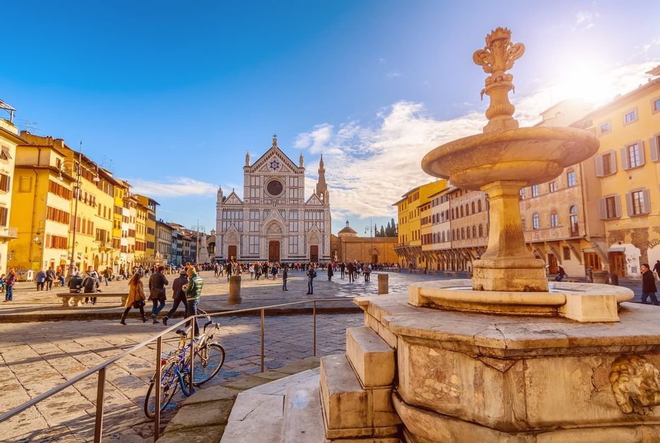 A stone fountain in the foreground of a sunny European square, with people walking in front of an ornate white basilica.