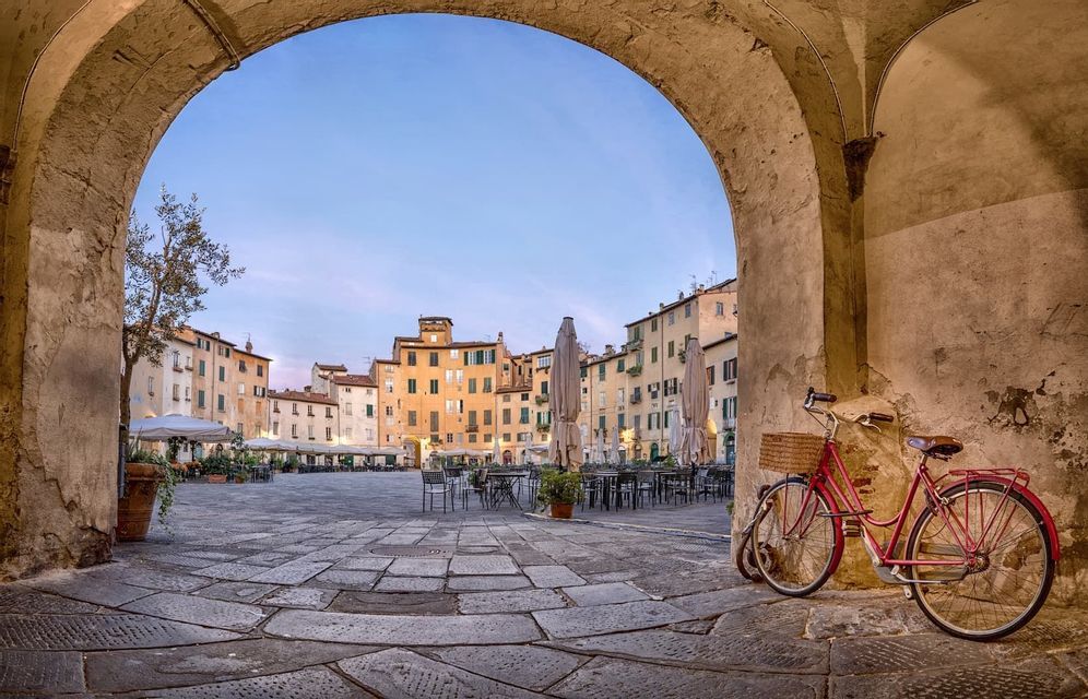 A red bicycle with a basket is parked under a stone archway, looking out onto a historic cobblestone plaza at dusk.