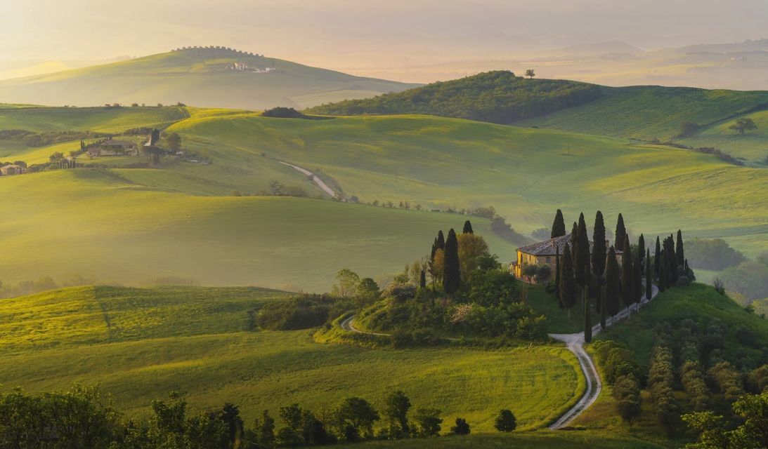 A villa surrounded by cypress trees sits on a rolling green hill amidst a hazy landscape at sunrise.