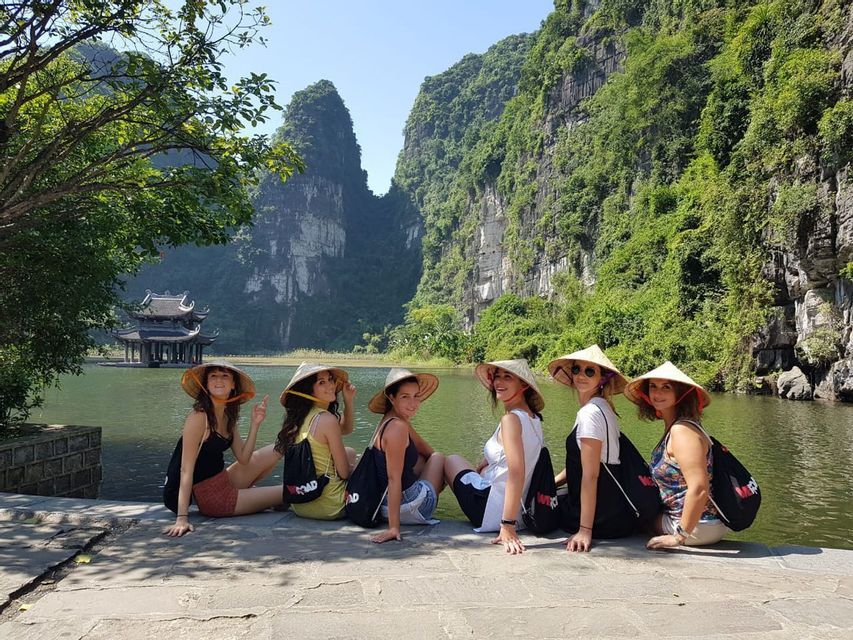 Eine WeRoad Gruppenreise mit sechs Frauen, die kegelförmige Hüte tragen und an einem See vor einer Kulisse aus grünen Bergen und einem Pavillon sitzen.