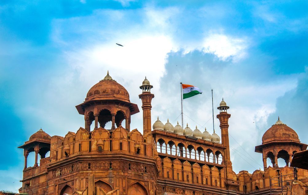 La bandera india ondea sobre una fortaleza ornamentada de arenisca roja con múltiples cúpulas y torres contra un cielo azul nublado.