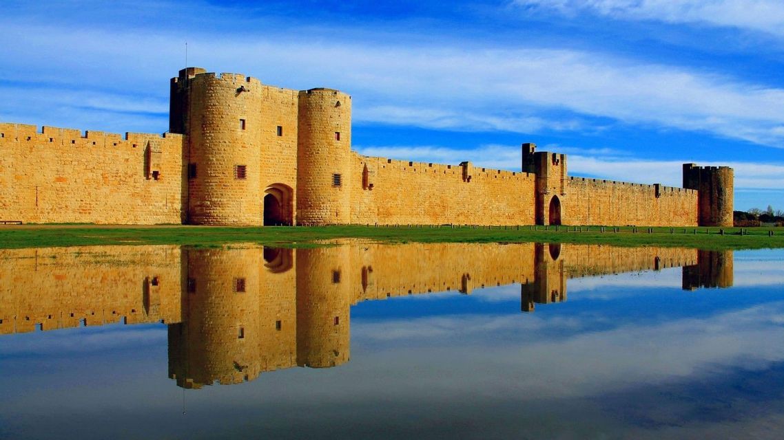 Una fortezza di pietra con torri rotonde e mura merlate si riflette in acque tranquille sotto un cielo azzurro brillante.