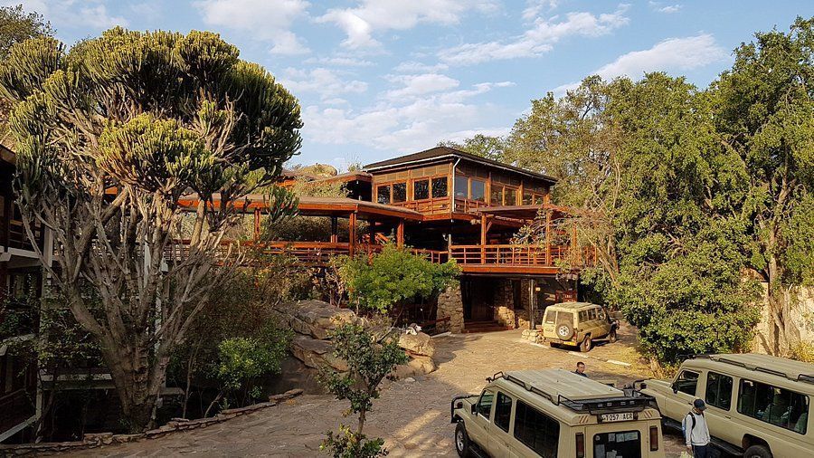 Safari-Jeeps stehen vor einer mehrstöckigen Holz-Lodge, umgeben von üppigen Bäumen und Felsen unter einem teilweise bewölkten Himmel.