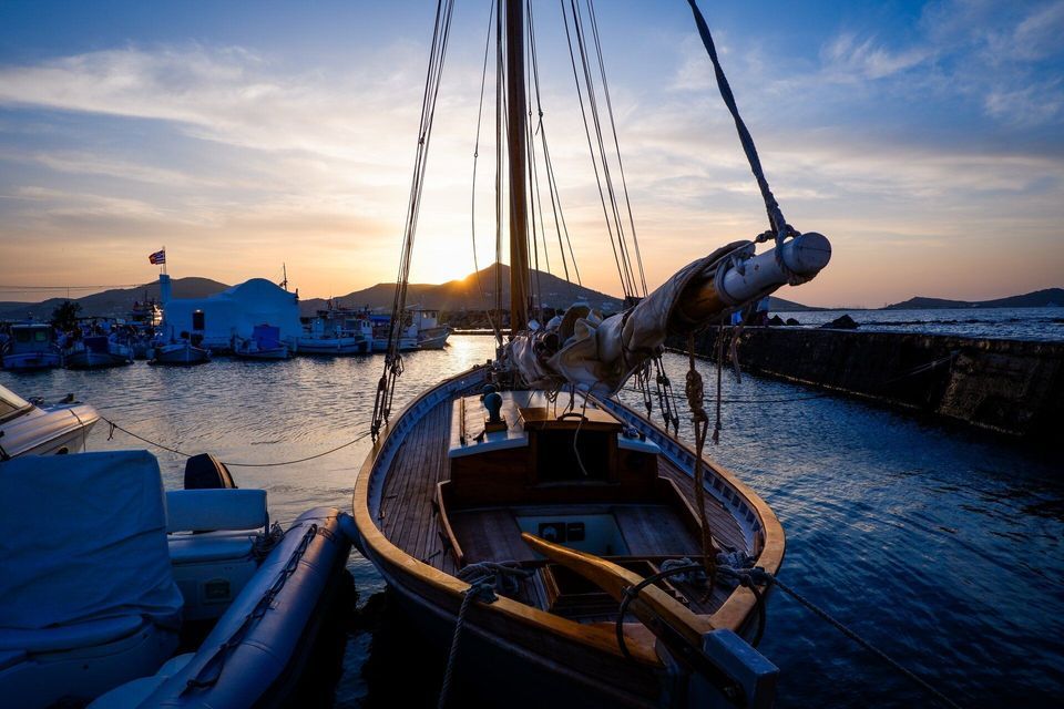 Una barca a vela in legno è ormeggiata in un porto al tramonto, con il sole che brilla dietro le colline distanti e altre barche nell'acqua.